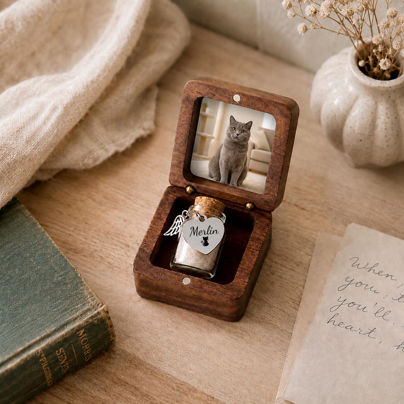 May include: A dark wood keepsake box with a hinged lid containing a photograph of a gray cat. Inside the box sits a small glass vial with a cork stopper, featuring a silver heart-shaped charm engraved with the name Merlin and a cat icon.