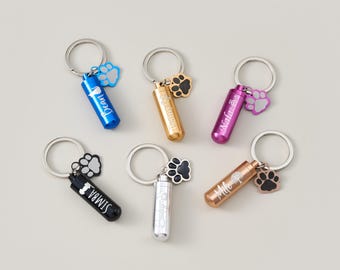 May include: A collection of pet memorial keychains in various colors, including blue, gold, pink, black, and silver. Each keychain features a small, cylindrical container with a name engraved on it, and a paw print charm.