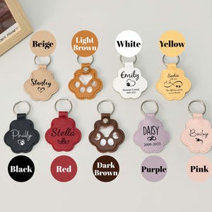 May include: A collection of personalized pet paw-shaped keychains in various colors, including beige, light brown, white, and yellow. Each keychain features a name and design, with a metal keyring attached. The image also shows keychains in black, red, dark brown, purple, and pink.