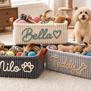 May include: Three rectangular storage baskets made of ribbed fabric with brown leather handles. The cream basket features the name Bella and a heart. The dark gray basket displays the name Milo and a paw print. The light gray basket shows the name Teddy and a bone.