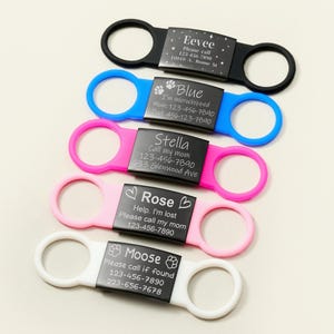Op de afbeelding: Vijf verschillende honden-ID-tags met gegraveerde namen en telefoonnummers. De tags zijn bevestigd aan kleurrijke siliconen halsbanden. De tags zijn zwart met witte gegraveerde tekst. De halsbanden zijn zwart, blauw, roze, wit en lichtroze.