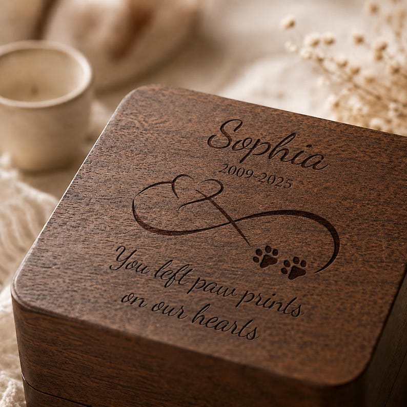 May include: A dark brown wooden box with an engraved infinity symbol, a heart, and two paw prints. The text reads: Sophia, 2009-2025, You left paw prints on our hearts. The lid features a smooth, grained texture and dark wood finish.