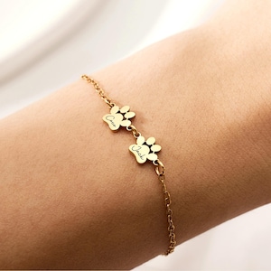 May include: Gold-coloured bracelet with two paw print charms. Each charm has a name engraved on it. The bracelet is worn on a person's wrist. The bracelet is a delicate piece of jewellery.