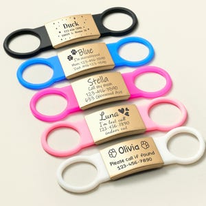 May include: Five colorful dog ID tags with engraved text and phone numbers. The tags are attached to different colored silicone collars. The text on the tags includes the dog's name, a message, and a phone number.