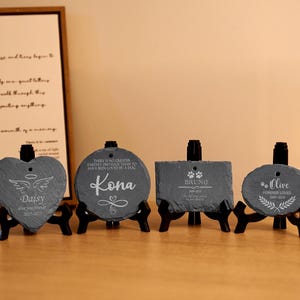 May include: Four slate pet memorial stones in various shapes: heart, circle, rectangle, and oval. Each stone is engraved with a pet's name, dates, and a loving message. The stones are displayed on black stands.