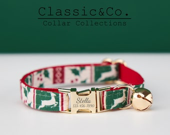 May include: A personalized cat collar with a festive design. The collar features a pattern of green holly and reindeer on a cream background, with red trim. A gold-colored buckle is engraved with the name "Stella" and a phone number. A gold bell hangs from the collar.