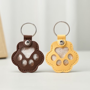 May include: Two paw-shaped keychains. One is brown, the other is yellow. Each has a silver keyring and a silver button. The paw pads are visible through the cutouts. The background is a light color.