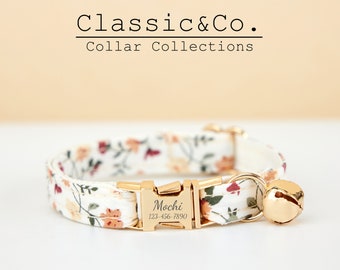 Könnte beinhalten: Ein weißes Katzenhalsband mit Blumenmuster und goldenen Beschlägen. Das Halsband hat eine Glocke und ein personalisiertes Namensschild mit der Aufschrift "Mochi 123-456-7890".