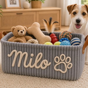 May include: A rectangular grey rope storage basket featuring the name Milo and a paw print design in cream-colored rope. The basket has tan leather handles and is filled with various dog toys, including a teddy bear, a bone, and balls.