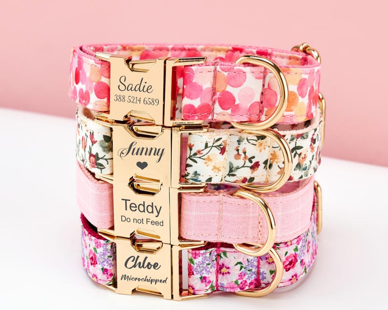 May include: Four personalized dog collars stacked. The top collar is pink with a polka dot pattern, the second has a floral design, the third is solid pink, and the bottom has a floral pattern. Each collar has a gold buckle with a name and contact information.