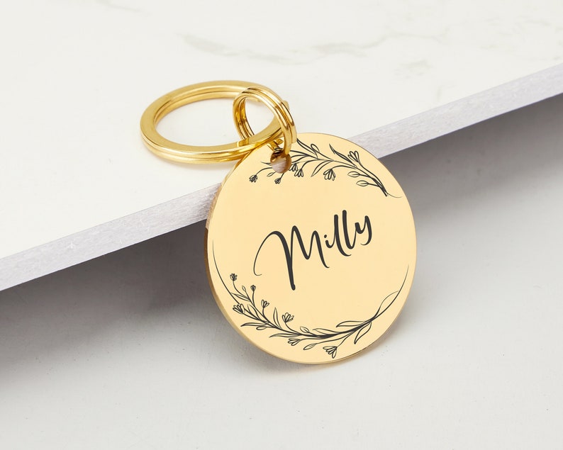 May include: Gold-colored metal pet tag with a floral design and the name "Milly" engraved on it.
