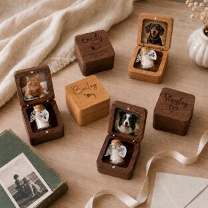 May include: Several square wooden keepsake boxes, some closed and some open, featuring engraved names like Emily, Bailey, and Stanley. Open boxes display pet photos and small glass vials with cork stoppers, each adorned with a heart-shaped tag.