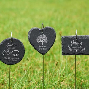 May include: Three black slate pet memorial garden stakes. One is round with the name "Sophia", dates, paw prints, and text. Another is heart-shaped with a tree design and the name "Steven" and dates. The third is rectangular with the name "Daisy", dates, and text.