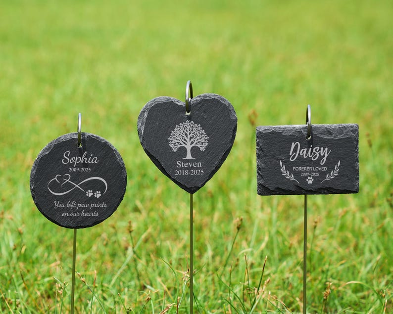May include: Three black slate pet memorial garden stakes. One is round with the name "Sophia", dates, paw prints, and text. Another is heart-shaped with a tree design and the name "Steven" and dates. The third is rectangular with the name "Daisy", dates, and text.