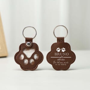 May include: Two brown leather paw print keychains with silver key rings. One keychain has a paw print cutout, the other has the name "Bruno" and the text "You left paw prints on our hearts."