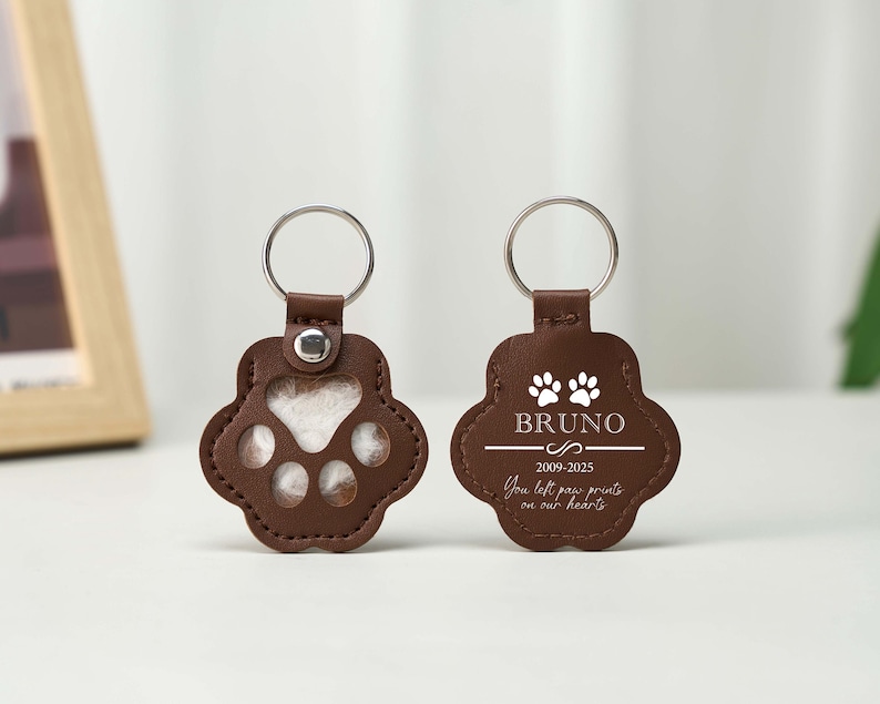 May include: Two brown leather paw print keychains with silver key rings. One keychain has a paw print cutout, the other has the name "Bruno" and the text "You left paw prints on our hearts."