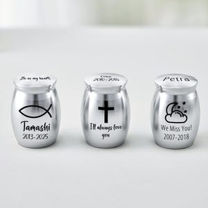 May include: Three silver memorial urns with black inscriptions. The urns feature a fish symbol, a cross, and a cloud with stars. Each urn has a name and dates. The text includes "Be in my heart", "I'll always love you", and "We Miss You!"