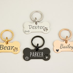 May include: Four personalized dog bone-shaped tags with key rings. The tags are in silver, gold, black, and rose gold. The names "Dexter", "Bear", "Parker", and "Bailey" are engraved on the tags.