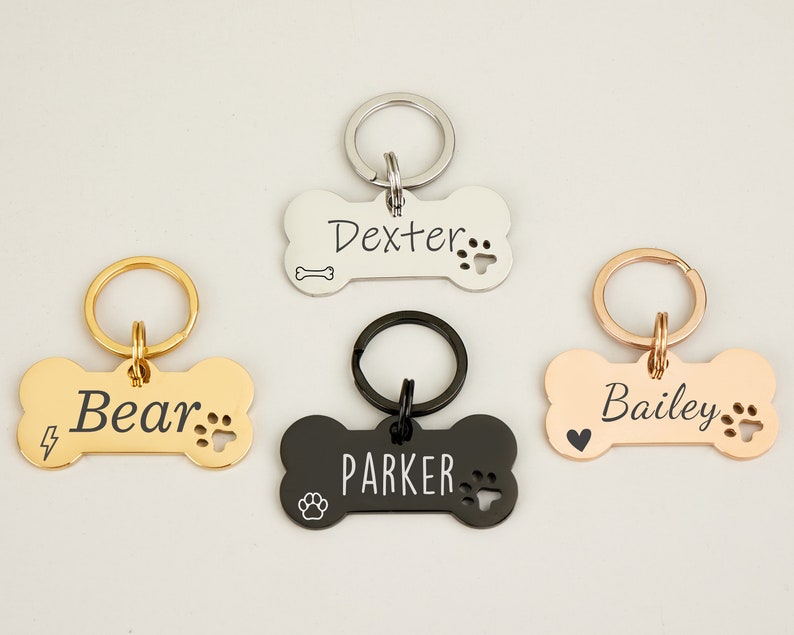May include: Four personalized dog bone-shaped tags with key rings. The tags are in silver, gold, black, and rose gold. The names "Dexter", "Bear", "Parker", and "Bailey" are engraved on the tags.