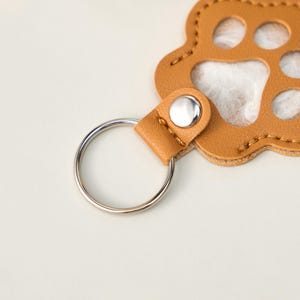 May include: A tan leather paw print keychain with a silver key ring. The keychain has a stitched border and a silver snap closure. The paw print design has cut-out circles for the paw pads.