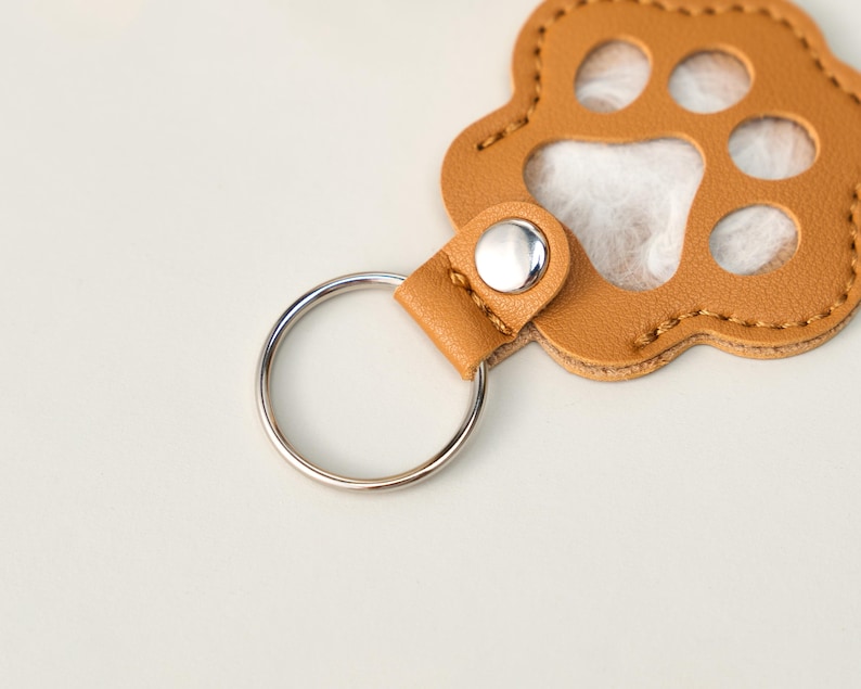 May include: A tan leather paw print keychain with a silver key ring. The keychain has a stitched border and a silver snap closure. The paw print design has cut-out circles for the paw pads.