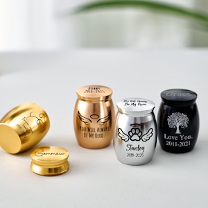 Op de afbeelding: Een verzameling kleine, decoratieve urnen in goud, brons, zilver en zwart. Elke urn heeft een deksel en is voorzien van gegraveerde tekst en ontwerpen, waaronder engelen vleugels, pootafdrukken en een boom. De urnen zijn voor gedenkdoeleinden.