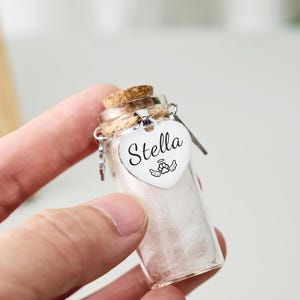 Może przedstawiać: Mała, przezroczysta szklana butelka z korkiem, wypełniona białą watą. Dołączona jest srebrna zawieszka w kształcie serca z imieniem "Stella" i skrzydłami anioła. Butelka jest trzymana w dłoni.