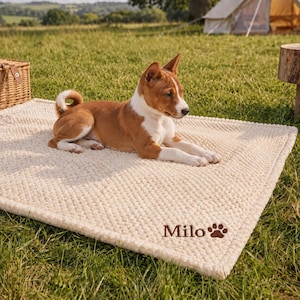 May include: A rectangular cream-colored textured pet mat featuring the name Milo embroidered in brown script alongside a brown paw print icon. A brown and white dog with pointed ears is lying down on the center of the soft, plush mat.