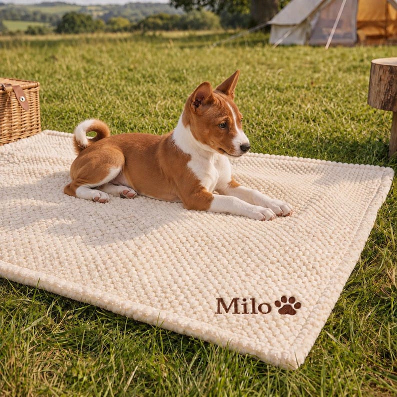May include: A rectangular cream-colored textured pet mat featuring the name Milo embroidered in brown script alongside a brown paw print icon. A brown and white dog with pointed ears is lying down on the center of the soft, plush mat.