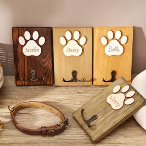 May include: Four rectangular wooden wall hooks featuring a light-colored wooden paw print cutout with the names Merlin, Daisy, Bella, and Vinny engraved. Each plaque includes a dark metal double hook. A brown leather pet collar is placed in the foreground.
