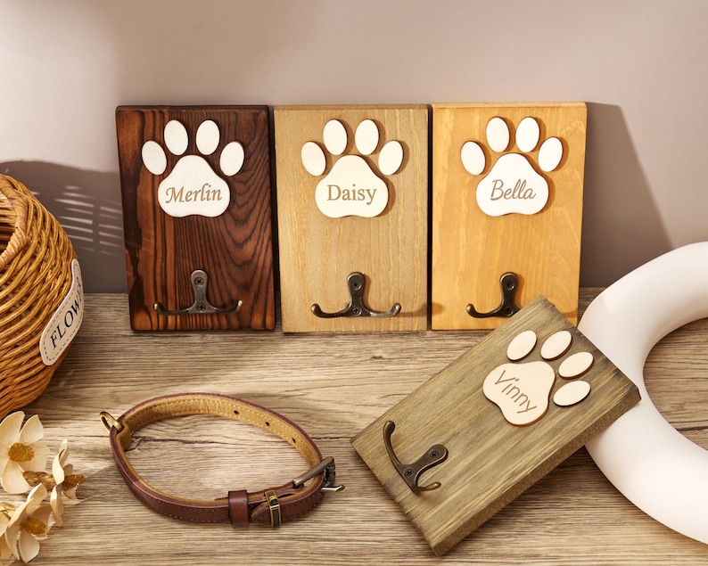May include: Four rectangular wooden wall hooks featuring a light-colored wooden paw print cutout with the names Merlin, Daisy, Bella, and Vinny engraved. Each plaque includes a dark metal double hook. A brown leather pet collar is placed in the foreground.