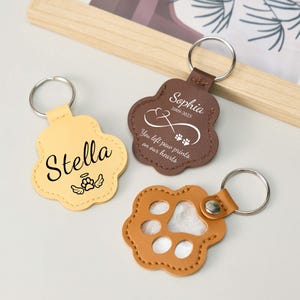 May include: Three paw-shaped keychains in tan, brown, and light orange. The tan keychain has the name "Stella" and an angel wing graphic. The brown keychain has the name "Sophia" and the text "You left paw prints on our hearts".