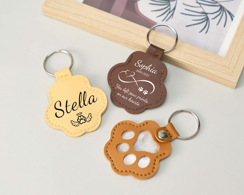 May include: Three paw-shaped keychains in tan, brown, and light orange. The tan keychain has the name "Stella" and an angel wing graphic. The brown keychain has the name "Sophia" and the text "You left paw prints on our hearts".