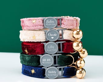 May include: A stack of five cat collars in various colors: pink, cream, red, green, and navy blue. Each collar has a gray buckle with a name and text, and a gold bell. The pink collar has gold stars, and the navy collar has gold stars.