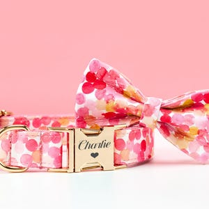 May include: A pink dog collar and matching bow tie set with a watercolor floral pattern. The collar features a gold-colored buckle engraved with the name "Charlie" and a heart symbol. The bow tie is pre-tied.