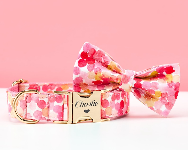 May include: A pink dog collar and matching bow tie set with a watercolor floral pattern. The collar features a gold-colored buckle engraved with the name "Charlie" and a heart symbol. The bow tie is pre-tied.