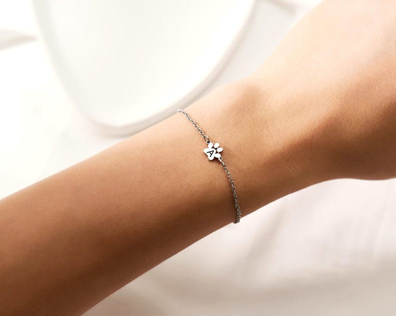 May include: A delicate silver bracelet featuring a paw print charm with the letter "A" in the center. The bracelet is worn on a wrist, showcasing its fine chain and minimalist design. The charm is small and detailed, adding a touch of charm.