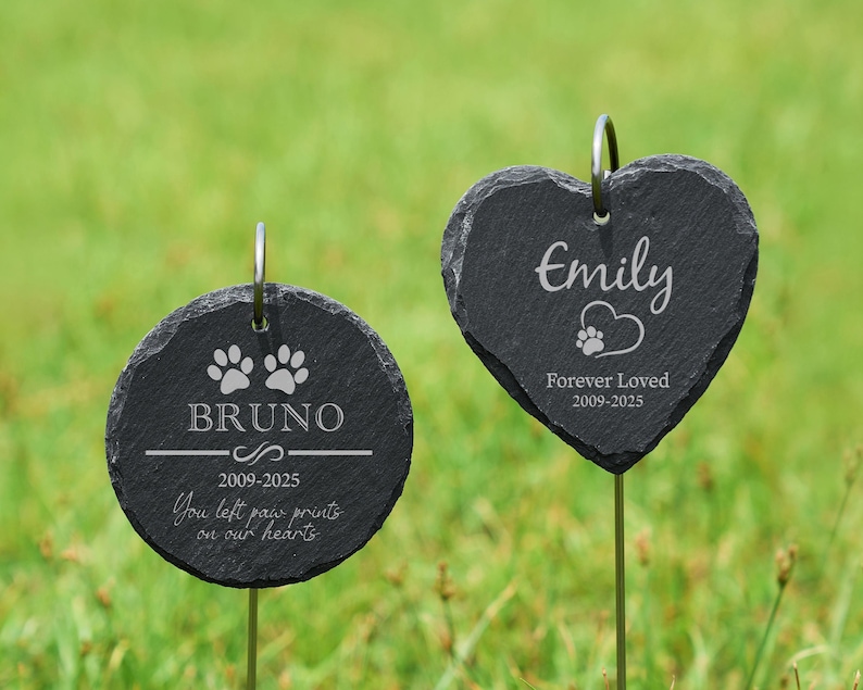 May include: Two black slate pet memorial garden stakes. One is round with the name "Bruno", paw prints, and the text "You left paw prints on our hearts". The other is heart-shaped with the name "Emily" and the text "Forever Loved".