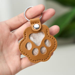 Peut inclure: Un porte-clés en forme de patte de chien en cuir marron avec un anneau argenté. Le porte-clés a un centre de couleur claire et est tenu dans une main. Le motif de la patte a trois découpes circulaires et une bordure cousue.