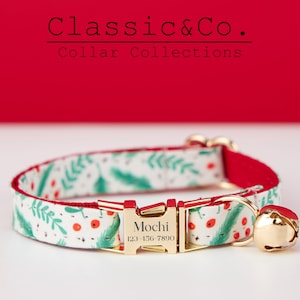 Puede incluir: Un collar de gato blanco con un patrón floral verde y rojo, una hebilla dorada con el nombre "Mochi" y el número de teléfono "123-156-7890" grabado en él, y una campanilla dorada.