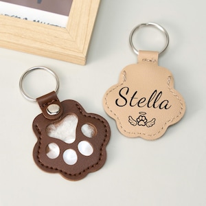 May include: Two paw-shaped keychains. One is brown with a clear center, the other is tan with the name "Stella" and a paw print with wings. Both have silver key rings. The keychains are likely pet-themed.