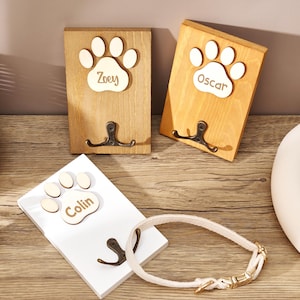 May include: Three rectangular wooden wall hooks featuring a light-colored wooden paw print cutout at the top. Each paw print is engraved with a name: Zoey, Oscar, or Colin. Below each paw print is a dark metal double hook for hanging items.
