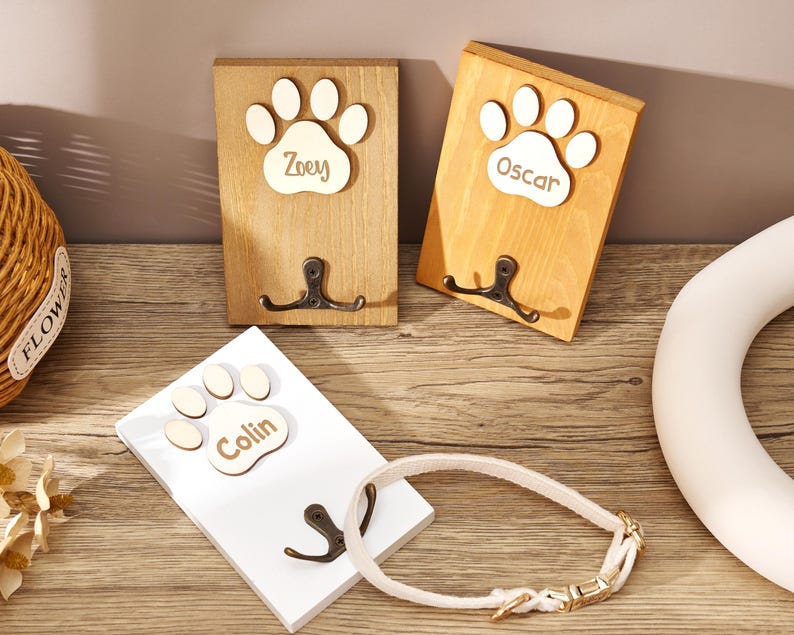 May include: Three rectangular wooden wall hooks featuring a light-colored wooden paw print cutout at the top. Each paw print is engraved with a name: Zoey, Oscar, or Colin. Below each paw print is a dark metal double hook for hanging items.