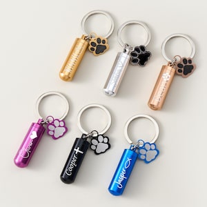 May include: Six keyrings with cylindrical pendants in gold, silver, rose gold, purple, black, and blue. Each keyring features a paw print charm and personalized text. The keyrings are designed for pet owners.