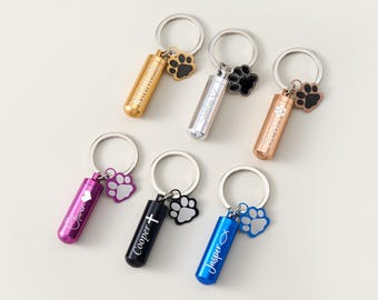 May include: A collection of six keychains with cylindrical pendants in gold, silver, rose gold, purple, black, and blue. Each keychain has a paw print charm and personalized text. The keychains are designed for pet owners.