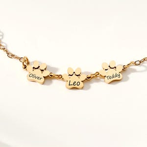 May include: A gold-colored necklace featuring three paw-shaped charms. Each charm has a name engraved: "Oliver", "Leo", and "Teddy". The necklace has a delicate chain and is likely made of stainless steel or a similar metal.