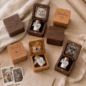 May include: A collection of square wooden keepsake boxes, some closed and some open, featuring engraved names like Sophia, Stanley, Bola, and Emily. Open boxes display pet photos and small glass vials with heart-shaped tags labeled Oscar, Annie, and Ruby.