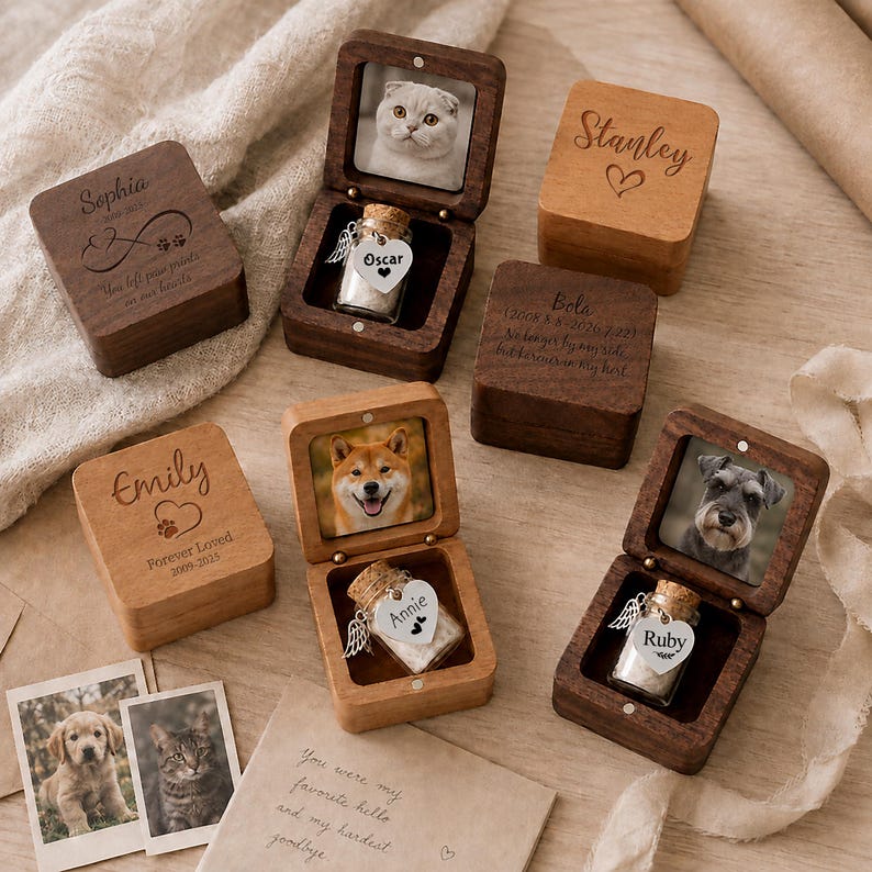 May include: A collection of square wooden keepsake boxes, some closed and some open, featuring engraved names like Sophia, Stanley, Bola, and Emily. Open boxes display pet photos and small glass vials with heart-shaped tags labeled Oscar, Annie, and Ruby.