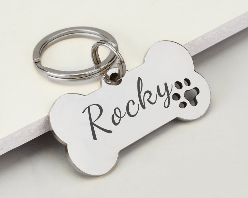 May include: Silver bone-shaped dog tag with a split ring. The tag is engraved with the name "Rocky" and a paw print.