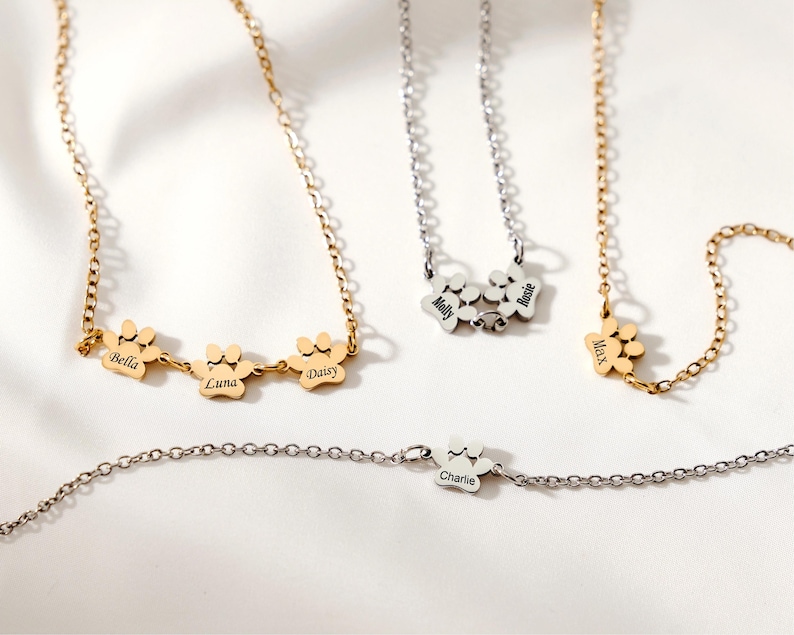 May include: A collection of personalized paw print necklaces and a bracelet. The necklaces are gold and silver-toned, featuring names like "Bella," "Luna," "Daisy," "Molly," "Rosie," and "Max." The bracelet has the name "Charlie."